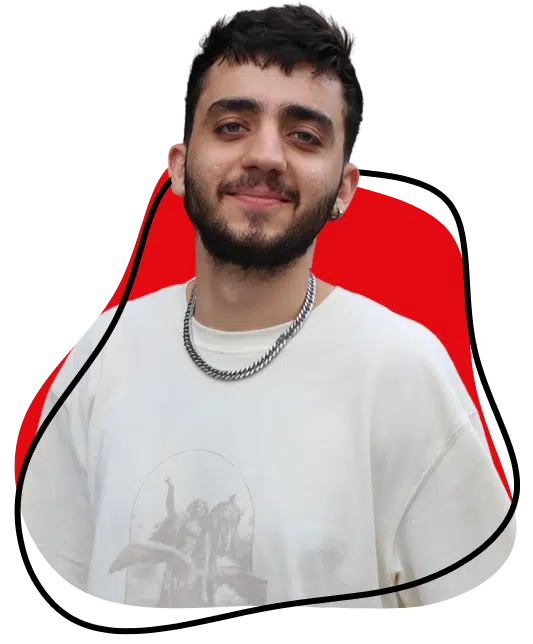 A young man with short dark hair and a beard smiles at the camera. He wears a white T-shirt with a graphic print and a silver chain necklace. The background features red and black abstract shapes.