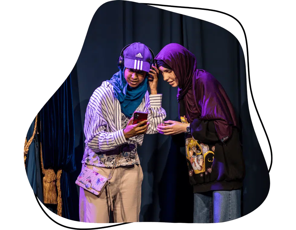 Two people wearing hijabs and casual clothing stand closely together on stage, sharing earphones and looking at a mobile phone, with a blue curtain as the background.