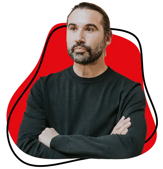 A man with long dark hair tied back and a beard stands with arms crossed, wearing a black jumper. The background features a red abstract shape outlined in black.