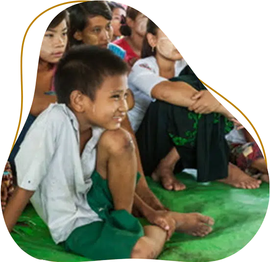 A group of children sit closely together on a green mat, with one smiling boy in the foreground wearing a white shirt and green shorts. They appear to be attentively watching something off-camera.