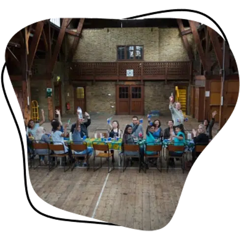 A group of people sit around a long table in a large hall with wooden floors and high ceilings, raising their hands and smiling at the camera. The hall has exposed beams and a rustic, spacious interior.