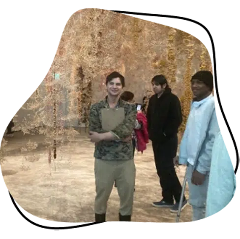 A group of people stand together in an indoor space with hanging decorations made of delicate, light-coloured materials. One person in front smiles with arms crossed whilst others chat in the background.