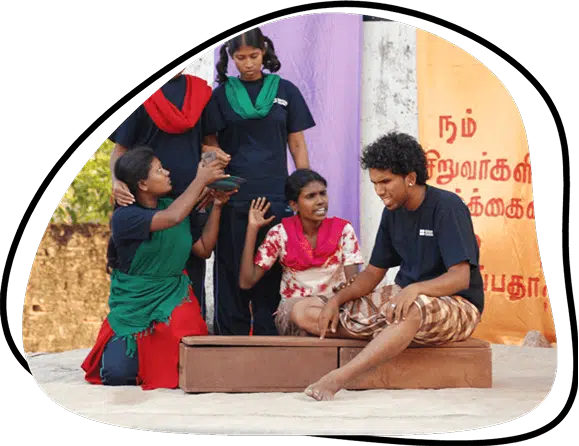 Five young people perform a dramatic scene outdoors. One person sits on a box, gesturing passionately, whilst others stand or kneel around, interacting with expressions of concern. A banner with Tamil writing is visible in the background.