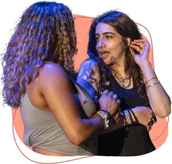 Two women standing close together, engaged in an animated conversation. One woman is touching her ear and gesturing, whilst the other faces her, listening attentively. Both have long hair and are wearing casual tops and jewellery.