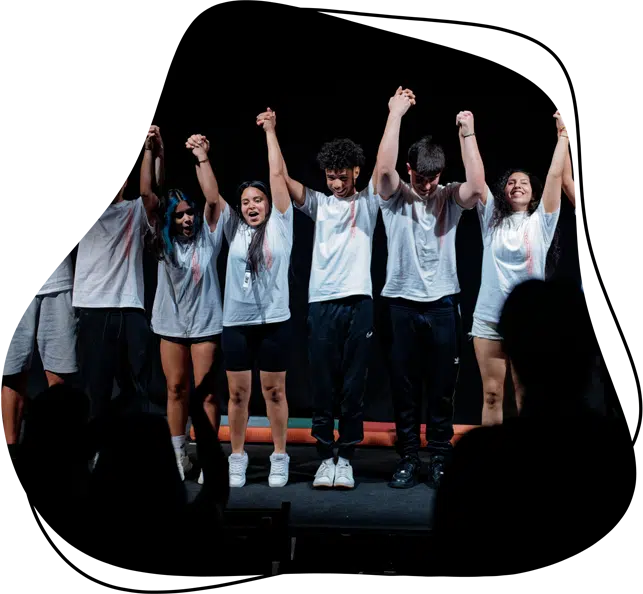 Six young people stand on stage holding hands and raising them in the air, smiling and wearing casual white shirts and dark trousers, while an audience in the foreground applauds.