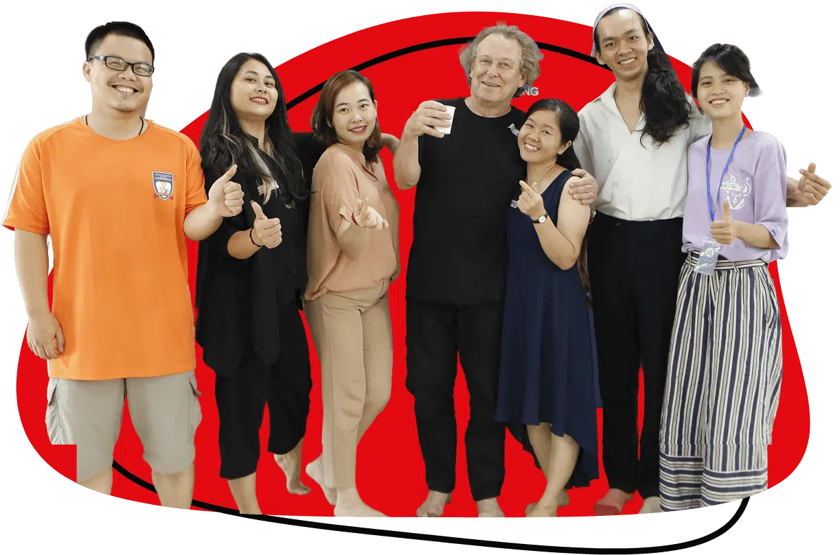 Seven people stand together smiling in front of a bold red and white background, with some giving thumbs up. The group includes both men and women, dressed casually and barefoot, appearing happy and friendly.