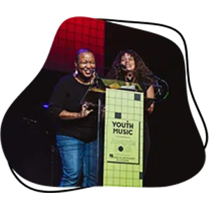 Two women smile whilst standing at a podium with a Youth Music sign, speaking into microphones on a stage with a red and black background.