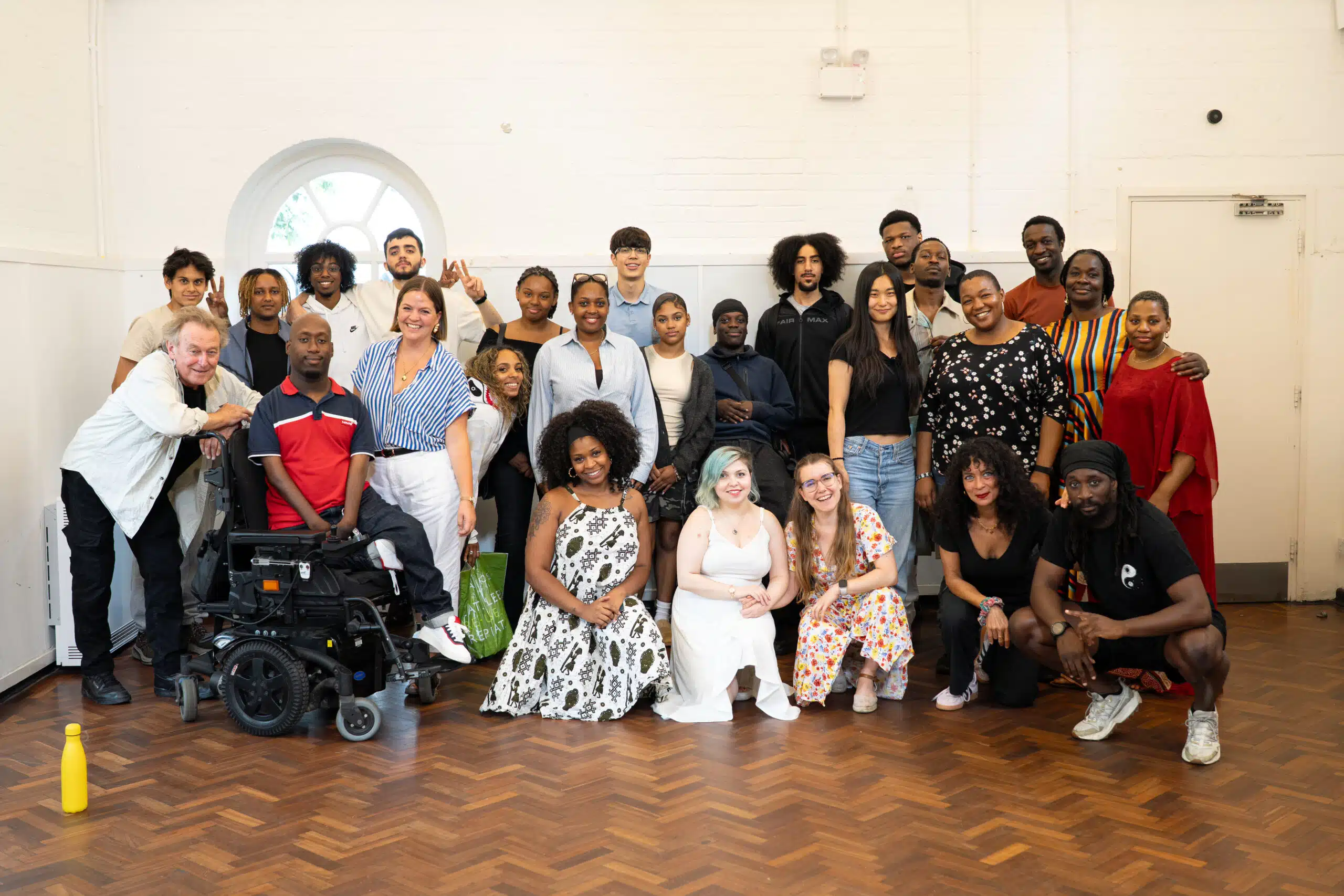 A diverse group of people, including individuals of different ages, races, and abilities, pose and smile together in a bright room with wooden floors and white walls. Some are seated whilst others stand behind them.