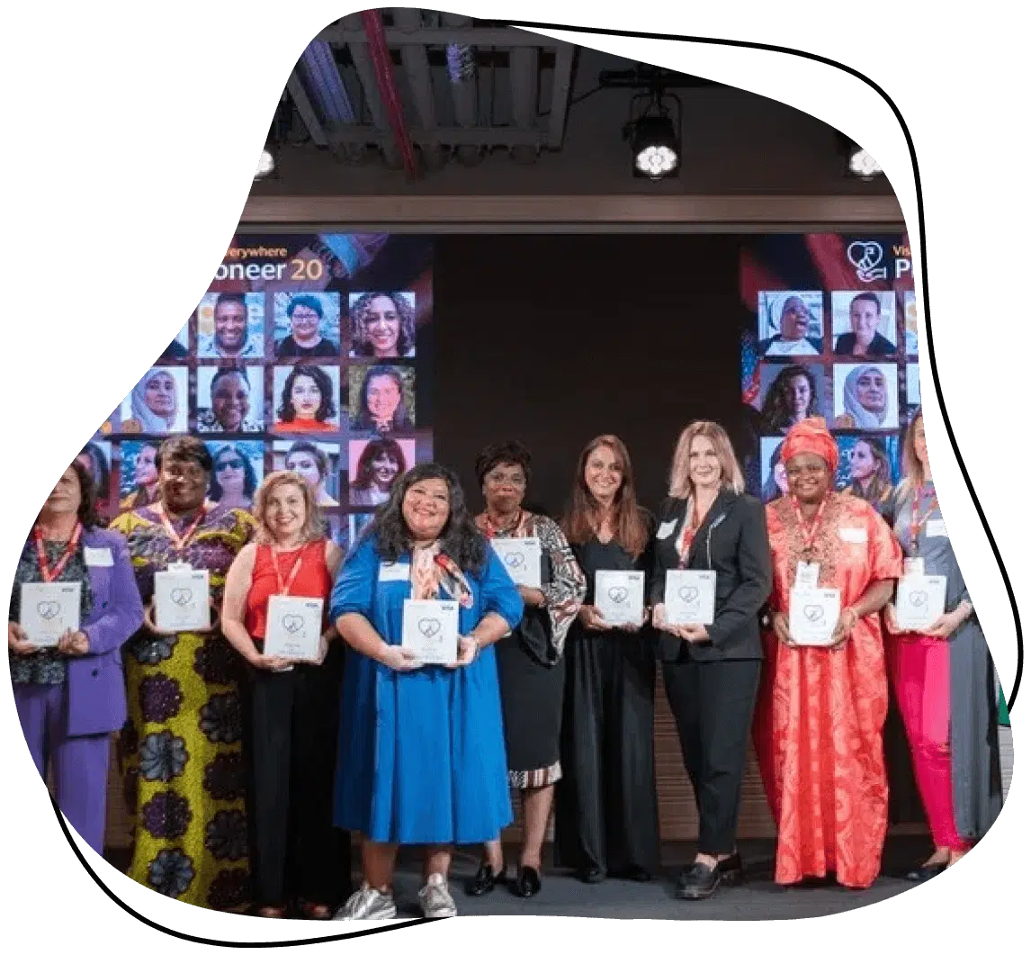 A diverse group of women stands on stage holding awards, smiling for a photo. Behind them, a screen displays many headshots and the text “Pioneer 20.” The setting appears to be a professional award event.