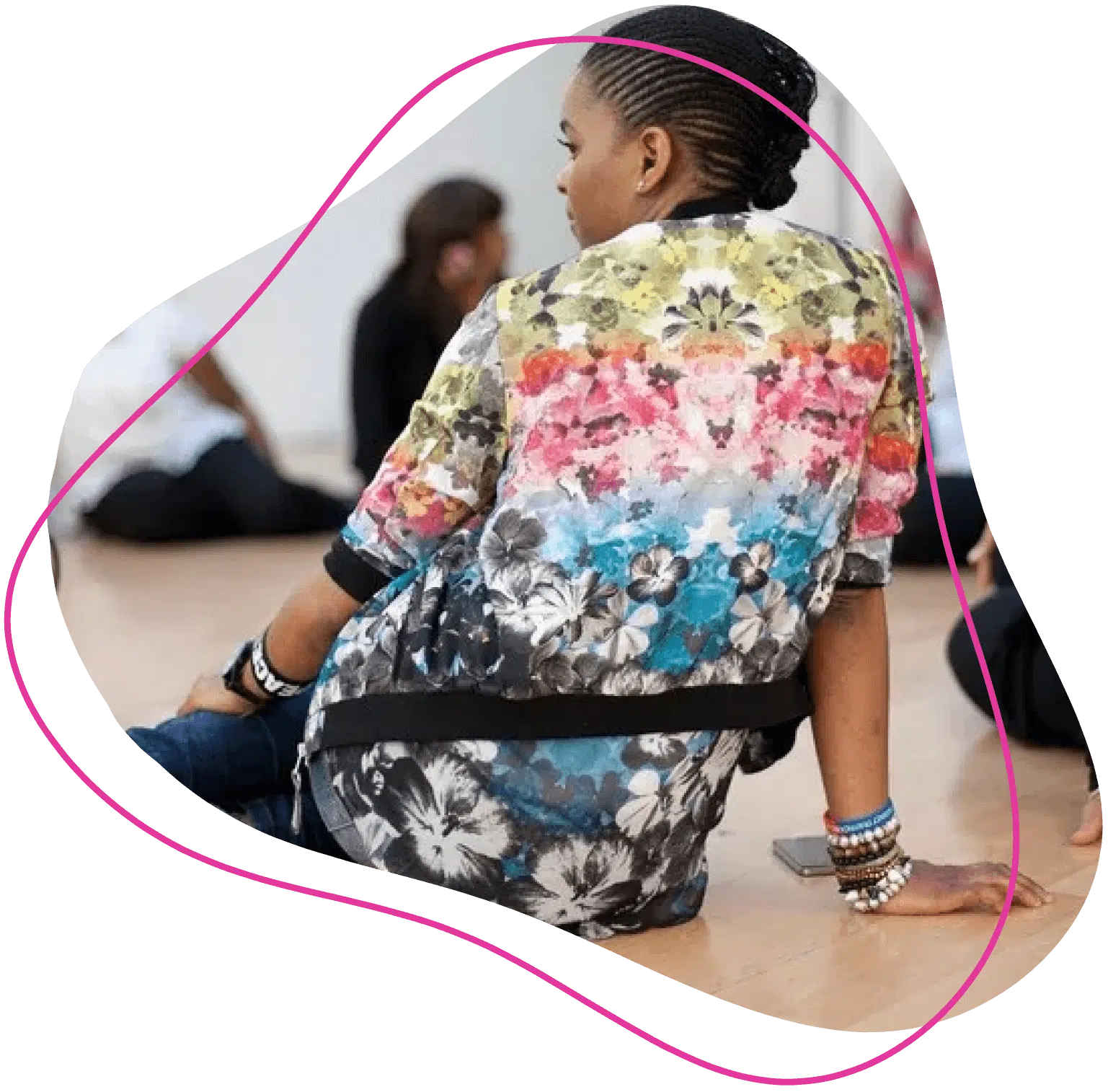 A person with plaited hair, wearing a colourful floral-patterned top and bracelets, sits on the floor with others in the background, outlined by a pink abstract shape.