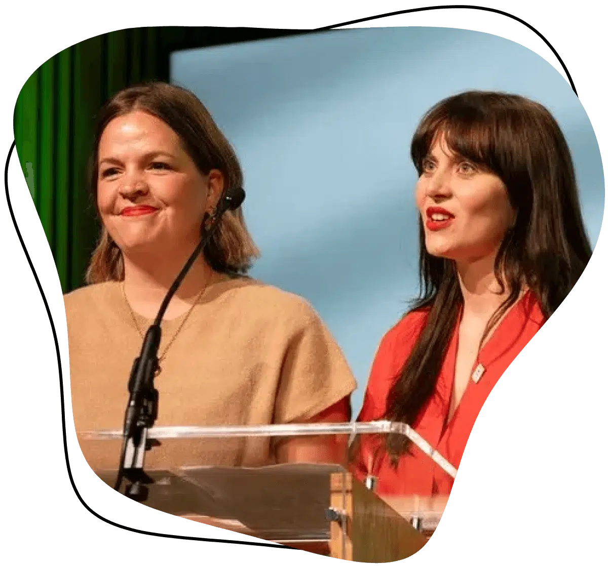 Two women standing behind a clear lectern with microphones, speaking or presenting on stage. One wears a tan outfit and the other wears red, both smiling. A blue screen and vertical green lines are in the background.