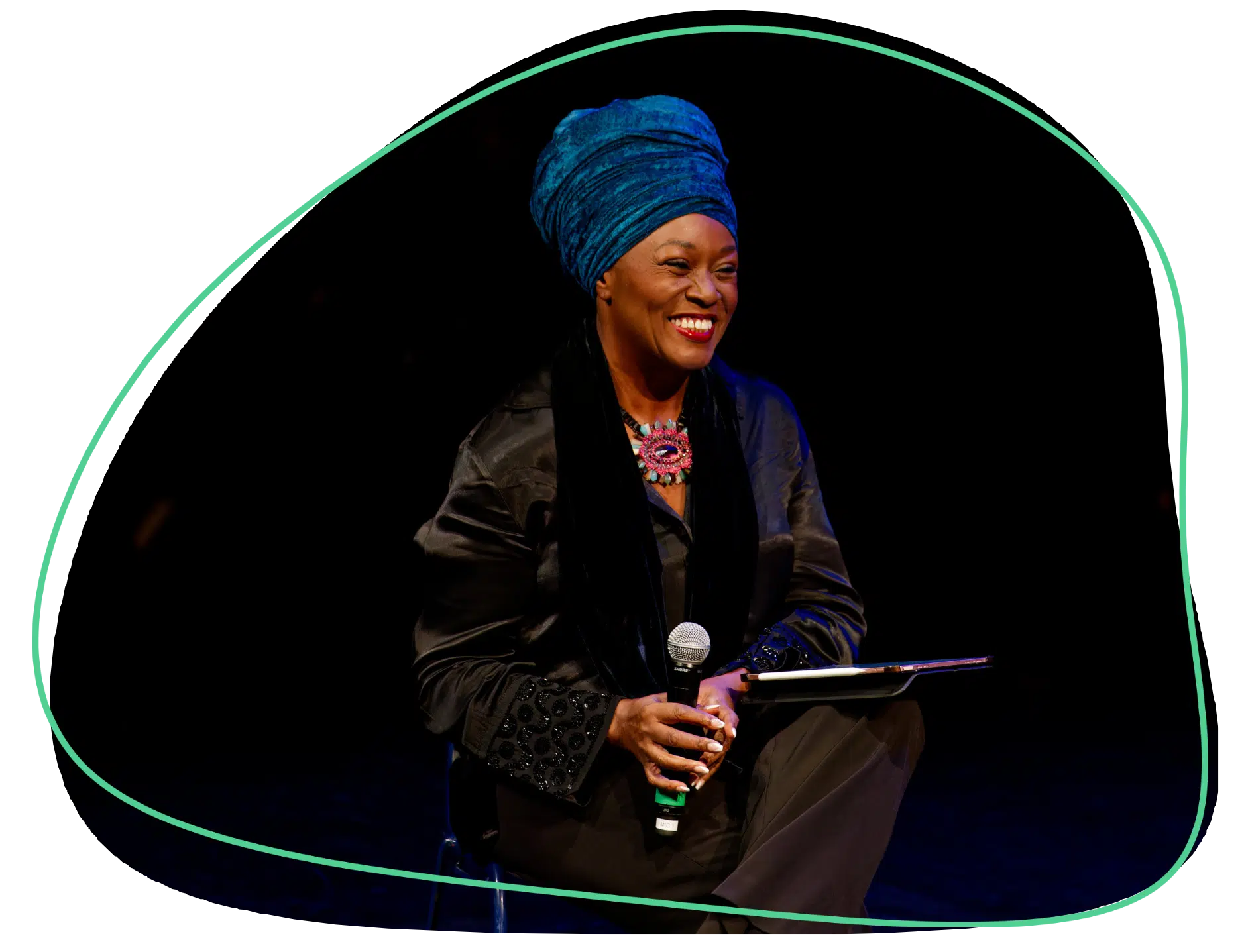A smiling woman wearing a bright blue headwrap and dark clothing sits onstage, holding a microphone and a tablet, with a decorative green line encircling her against a black background.