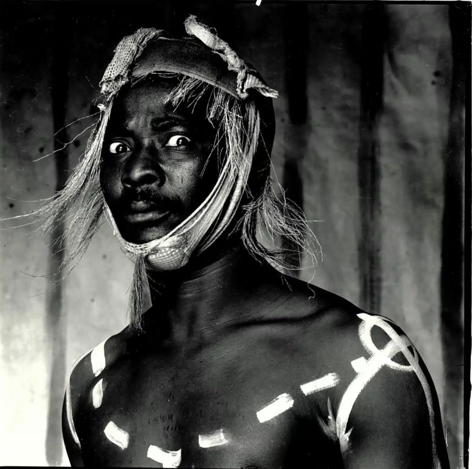 A person with wide eyes wears a headpiece with loose strands and has white painted lines and shapes on their chest and shoulder, standing against a plain background.
